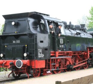 Eine alte schwarz-rote Dampflokomotive der Sauschwänzlebahn-Linie, mit zwei Personen im Führerstand, tuckert an einem bewölkten Tag umgeben von üppigem Grün entlang der Schienen und weckt ein Gefühl von Nostalgie.